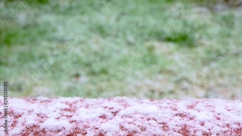 First snow falling onto red wooden railing on deck at extreme shallow depth of field in front of green grass on lawn in backyard 