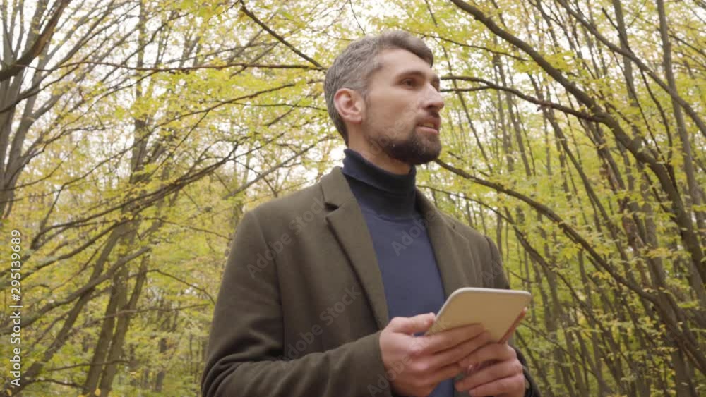 Handsome Caucasian man looking up and around in the forest and holding small tablet. Confident tourist enjoying quiet autumn day outdoors. Handsome Caucasian man looking up and around in the forest and holding small tablet. Confident tourist enjoying quiet autumn day outdoors.