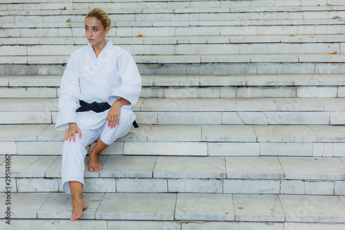 Young attractive woman in white kimono with black belt. Sport woman sitting on stairs outdoors. Martial arts