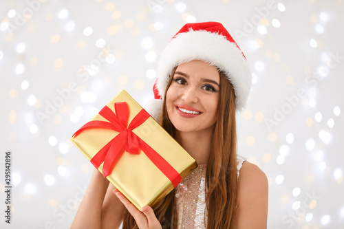 Happy young woman in Santa hat with gift box against blurred Christmas lights