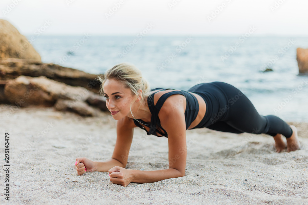 Young pretty sport blond woman in sportswear is practicing an exercise plank on the sand at wild beach. Outdoor training