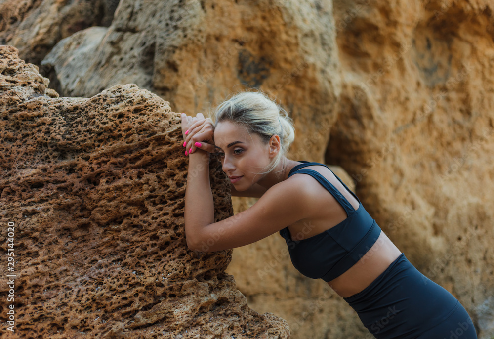 Fototapeta premium Portrait of a tired fitness woman in sportswear leaning on a stone on a wild beach. Outdoor training concept