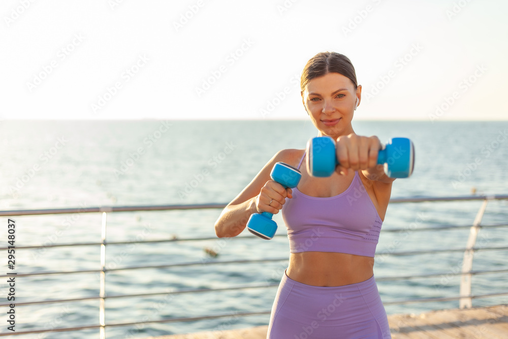 Young fit woman practices punches with dumbbells on the beach at sunrise. Morning workout with free weights Young fit woman practices punches with dumbbells on the beach at sunrise. Morning workout with free weights