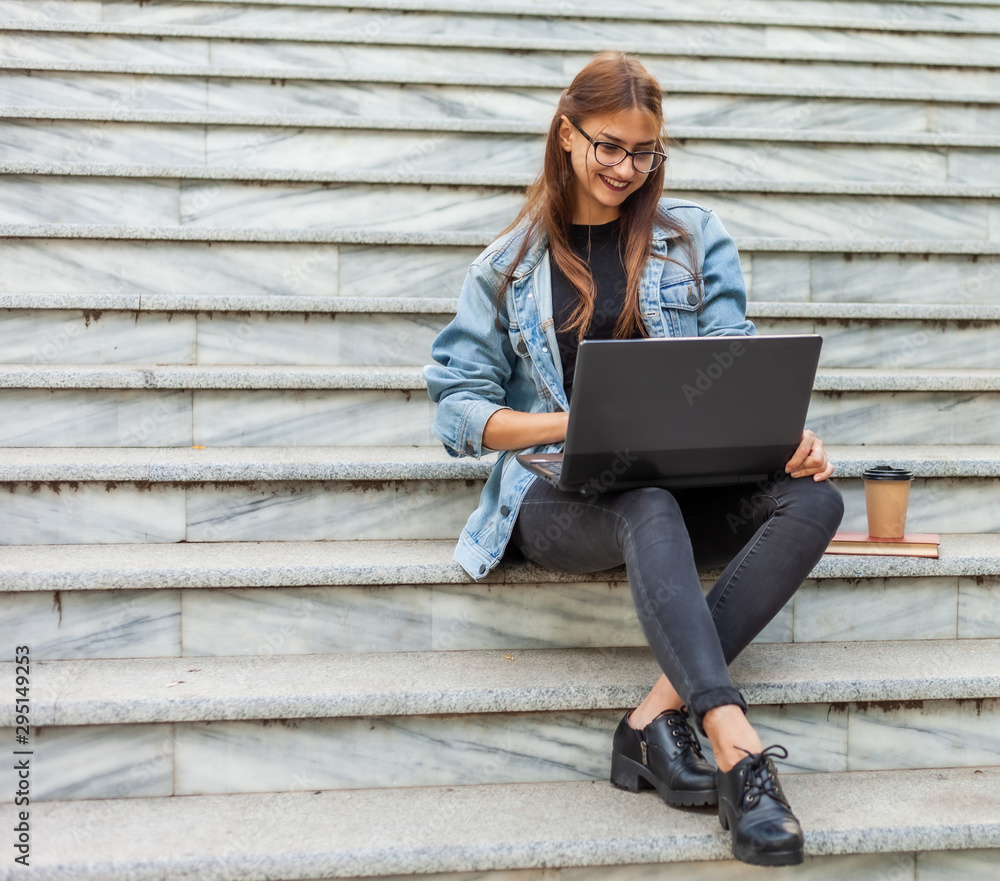 Young modern woman student in a denim jacket sitting on the stairs and watching video on laptop. Distance learning. Modern youth concept. Young modern woman student in a denim jacket sitting on the stairs and watching video on laptop. Distance learning. Modern youth concept.