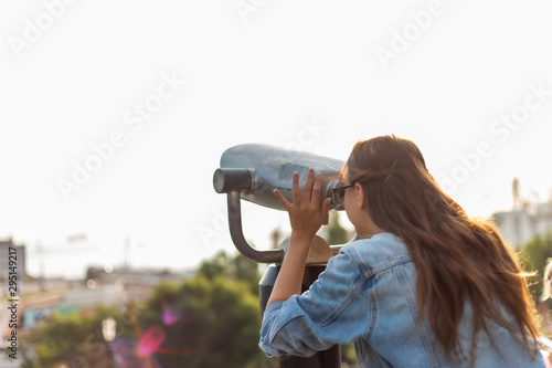 Young woman tourist in a denim jacket and glasses looks in the city binoculars in the city. Travel concept