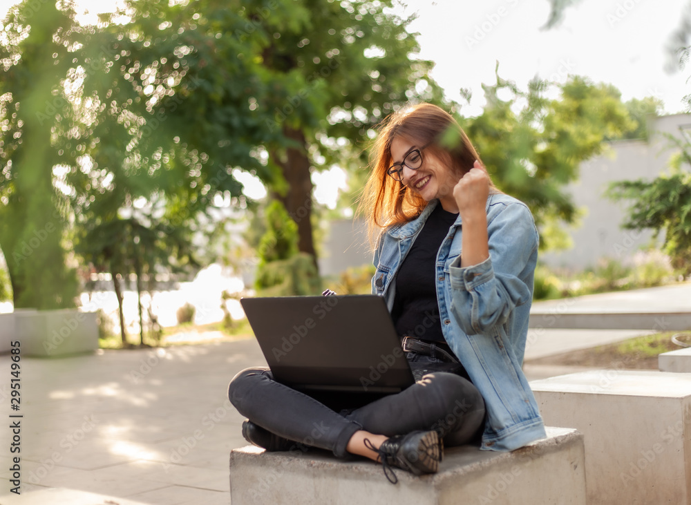 Young happy woman student in a denim jacket sitting at park and looks at the laptop screen. Distance learning. Online call. Modern youth concept. Young happy woman student in a denim jacket sitting at park and looks at the laptop screen. Distance learning. Online call. Modern youth concept.