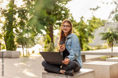 Young modern woman student in a denim jacket sitting at park and uses laptop with coffee cup on hand. Distance learning. Modern youth concept.