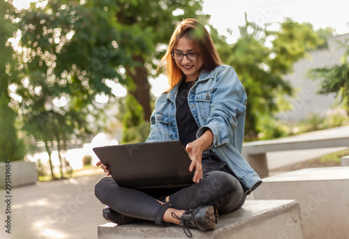 Young happy woman student in a denim jacket sitting at park and looks at the laptop screen. Distance learning. Online call. Modern youth concept.