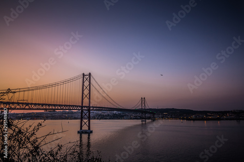 Fototapeta Naklejka Na Ścianę i Meble -  The 25 de April Bridge in Sunset Lights. Amazing view on Tagus River and one of the most famous and popular tourist sights in Lisbon. Portugal's beautiful landmark