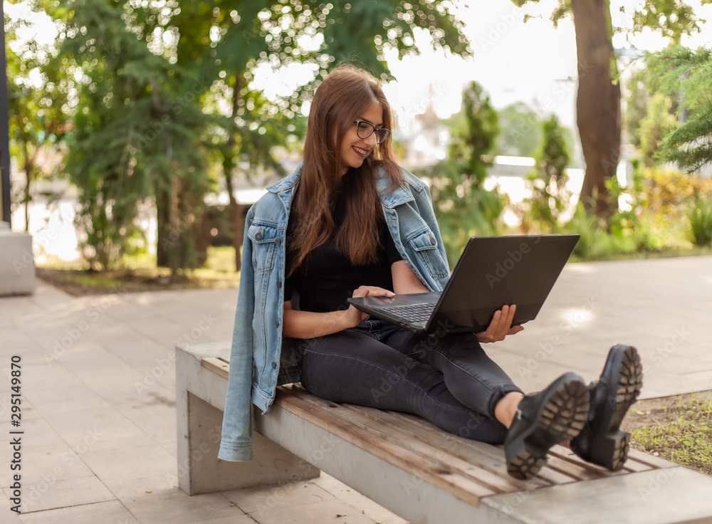 Online call. Distance learning. A young woman student in a denim jacket and glasses looks at a laptop screen while sitting on bench in park Online call. Distance learning. A young woman student in a denim jacket and glasses looks at a laptop screen while sitting on bench in park