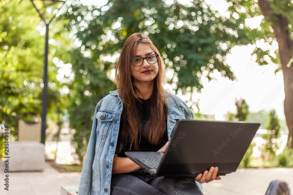 Distance learning. Young woman student in a denim jacket and glasses looks at camera whith aptop in hand while sitting on bench in park Distance learning. Young woman student in a denim jacket and glasses looks at camera whith aptop in hand while sitting on bench in park