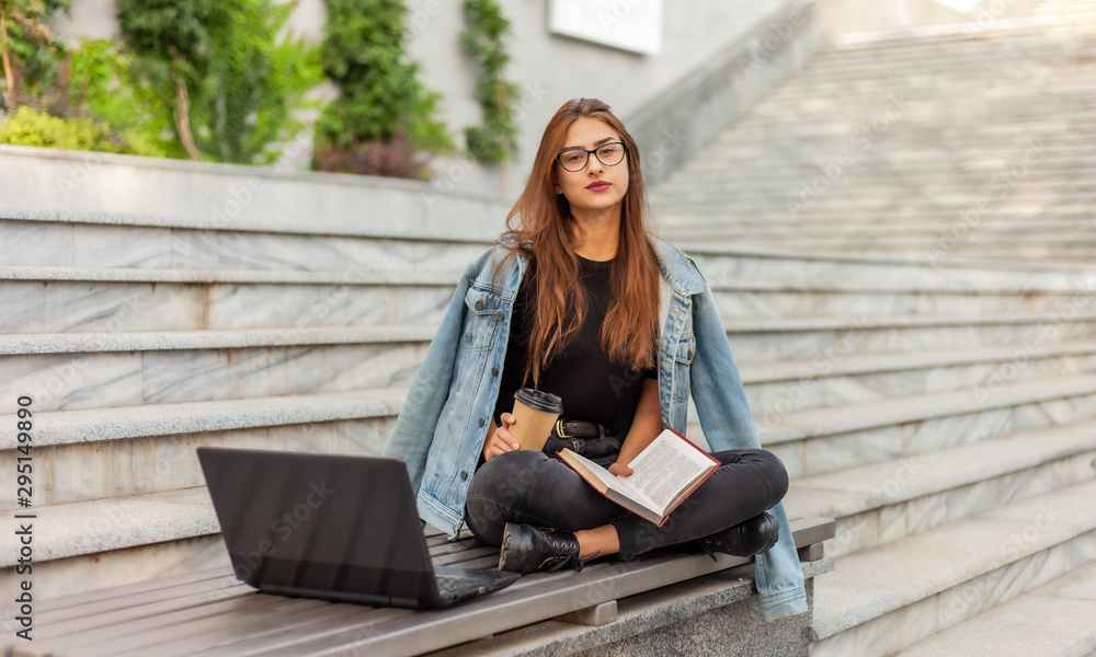 Obraz premium Modern students. Distance learning. Young hipster girl reads book while sitting on bench with a laptop