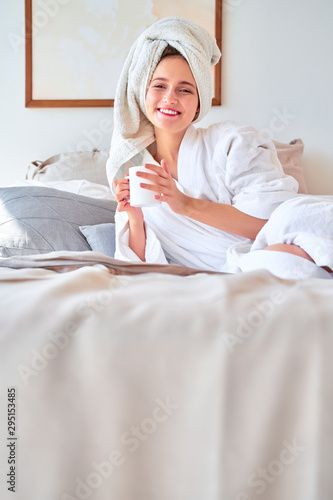 Photo of smiling woman in white bathrobe with mug of tea in her hands lying on bed.