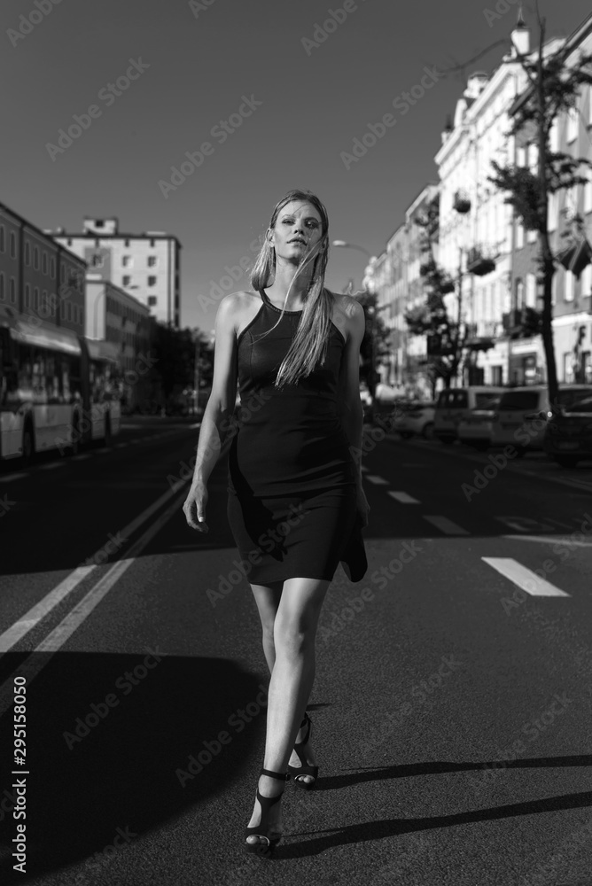 Vertical, black and white photo of elegant woman, in classic little black dress and high heels. Walking in the middle of the street with city buildings and cars in the background.