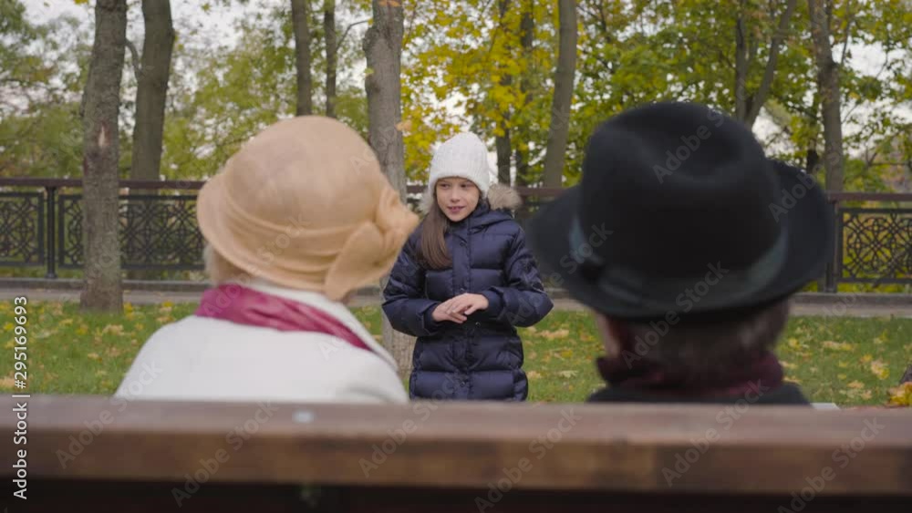 Portrait of a young cheerful Caucasian girl in white hat jumping like a rabbit in front of her grandparents. Happy family spending autumn day outdoors. Portrait of a young cheerful Caucasian girl in white hat jumping like a rabbit in front of her grandparents. Happy family spending autumn day outdoors.