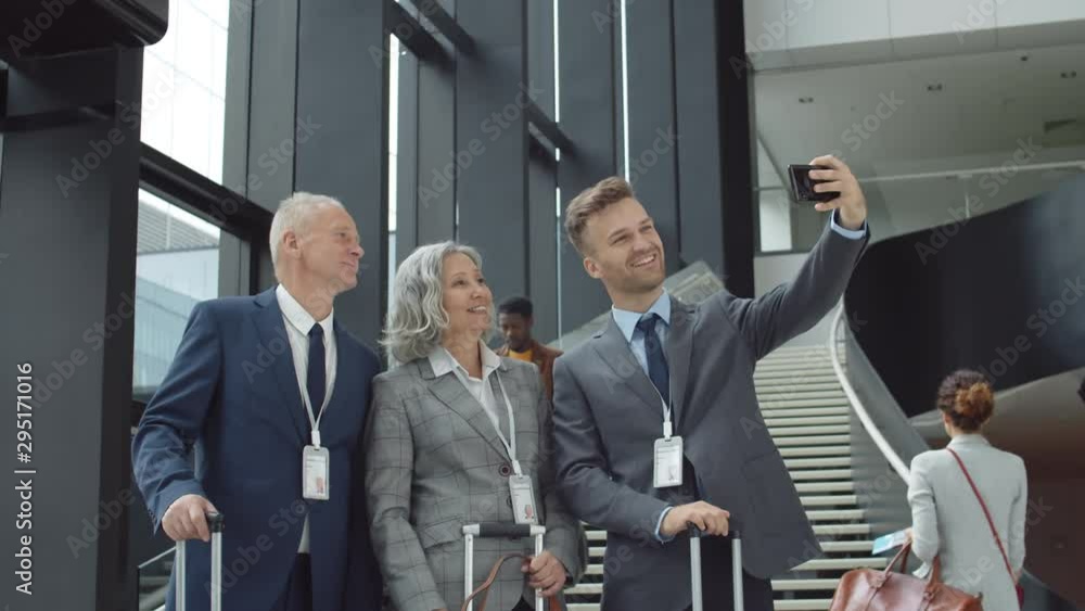 Tilt down of two Caucasian businessmen and businesswoman with suitcases in their hands standing in airport and making selfie using phone camera Tilt down of two Caucasian businessmen and businesswoman with suitcases in their hands standing in airport and making selfie using phone camera