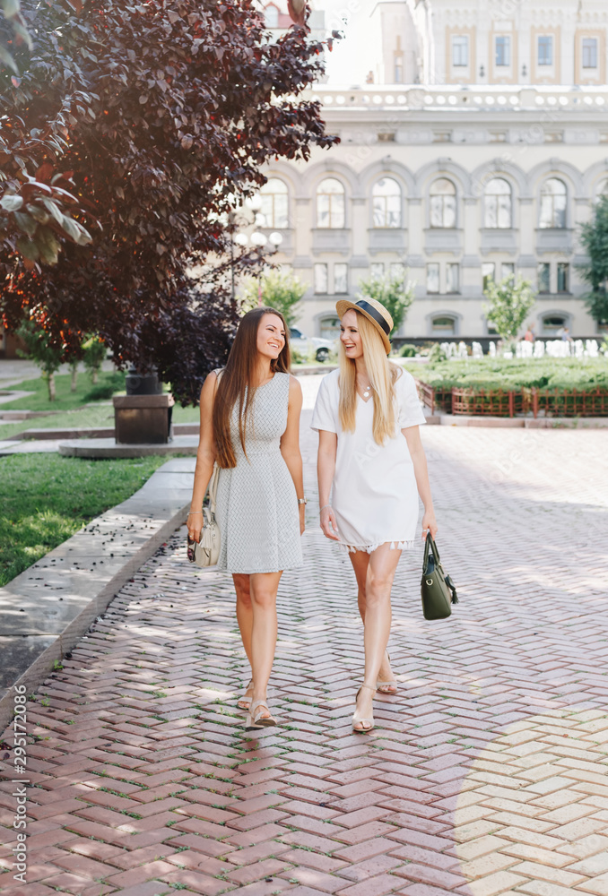 Two happy smiling girls are walking in the sunny city. Beautiful blonde and brunette walking down the street, students, travelers