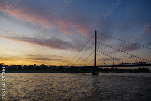 Silhouette view of city on riverside, Oberkasselerbrücke suspension bridge and Rhine River with beautiful dramatic purple, blue and golden sky during twilight sunset time.