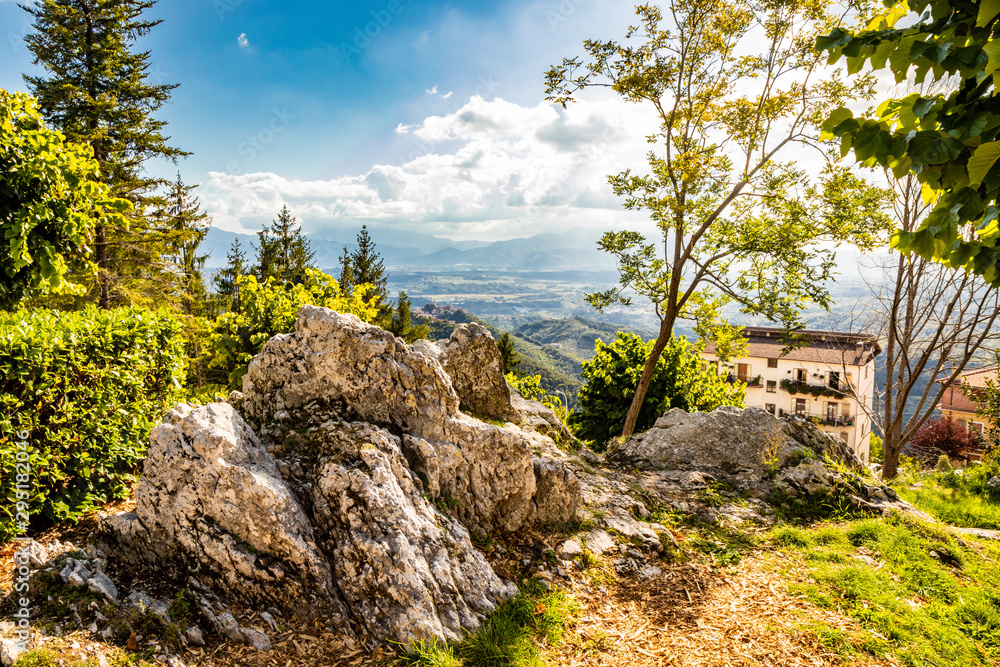 Bellegra, Rome, Lazio, Italy - The panorama seen from Bellegra. The cloudy blue sky. Mountains and sunbeams. Glimpse of a public garden, with trees and spikes of rock.
