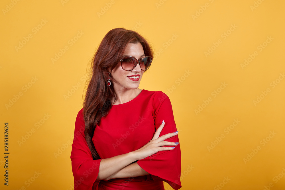 Fototapeta premium Young beautiful woman over isolated yellow background standing. Cheerful, smiling girl looking to the side. Lady in red clothes wearing brown sunglasses.