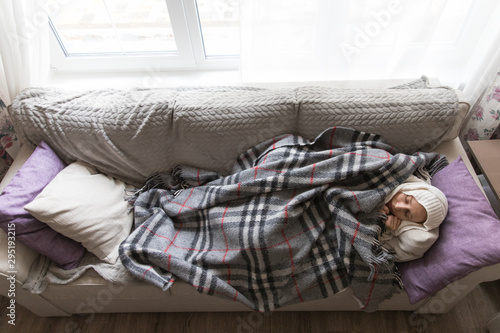 Sick young woman slleping, lying on sofa, wrapped in wool checkered plaid, tries to warm herself, having flu symptoms, feels cold, top view, wide angle. Cold, virus, sickness concept photo