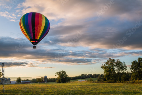 Rainbow hot-air balloon floats over farm field on a late summer morning as the sun rises
