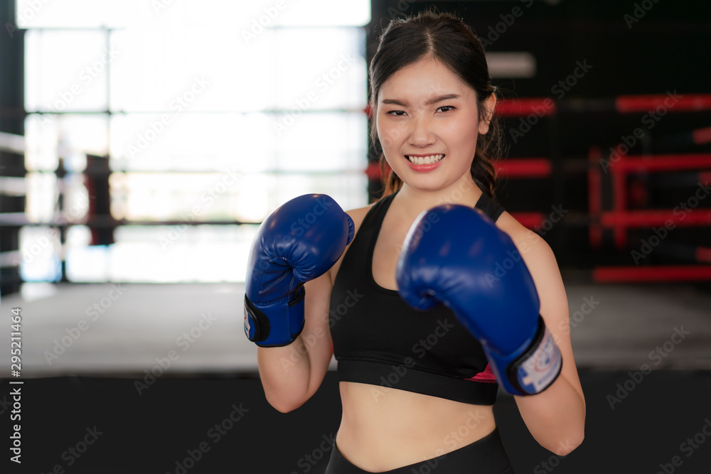  Young Asian Boxing fitness woman smiling happy wearing blue boxing gloves with Boxing Stadium in background. Portrait of sporty fit for healthy lifestyle Asian model of boxing gym concept..