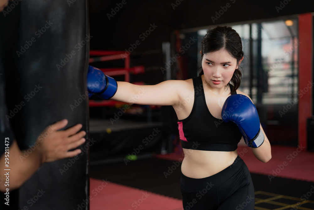 Portrait of Asian confident young boxer woman with blue boxing gloves, punching a Bag with trainer in professioal gym. Sporty fit for healthy lifestyle Asian model of boxing gym concept..
