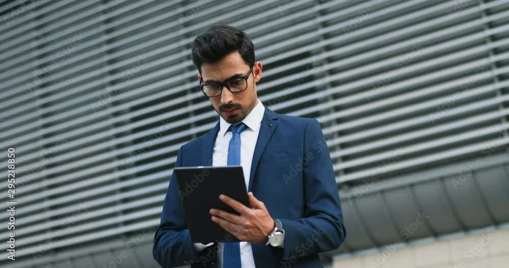 Arabian young handsome businessman in suit, tie and glasses working and tapping on the tablet computer while standing outside. Arabian young handsome businessman in suit, tie and glasses working and tapping on the tablet computer while standing outside.