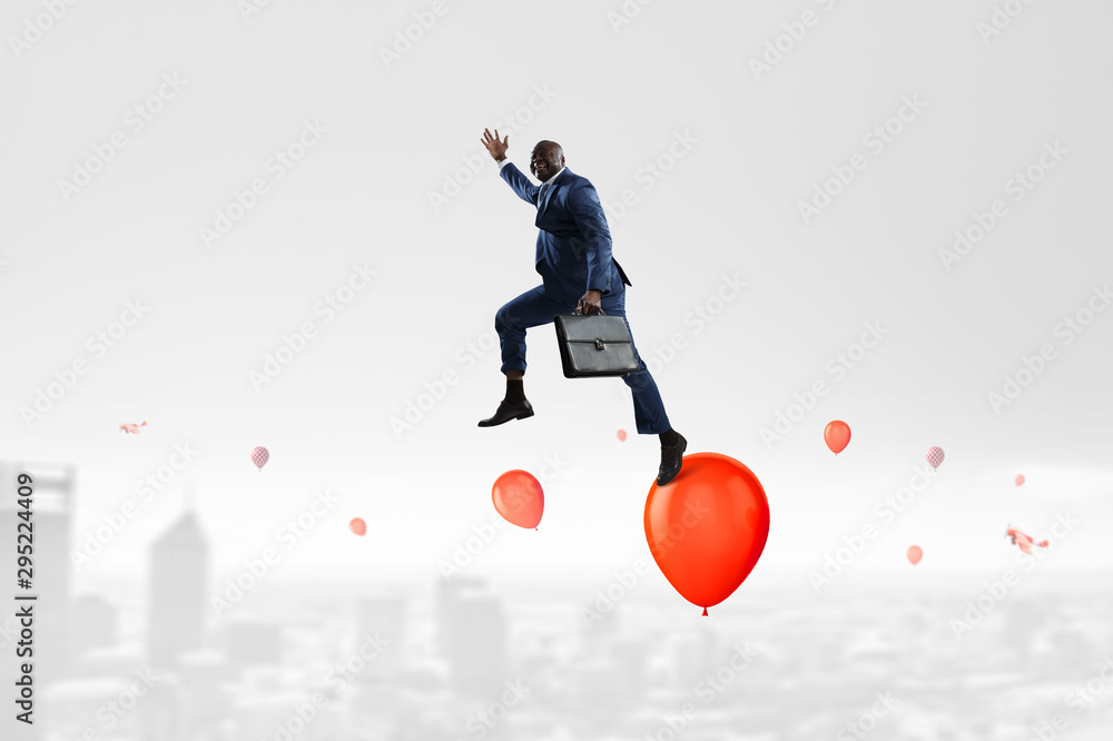 Black smiling businessman jumping over red ballons in the sky over urban area Black smiling businessman jumping over red ballons in the sky over urban area