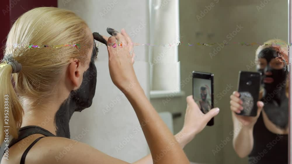 A woman in the bathroom near the mirror puts a black mud mask on her face and films herself on a smartphone camera, talking about the procedure. A woman in the bathroom near the mirror puts a black mud mask on her face and films herself on a smartphone camera, talking about the procedure.