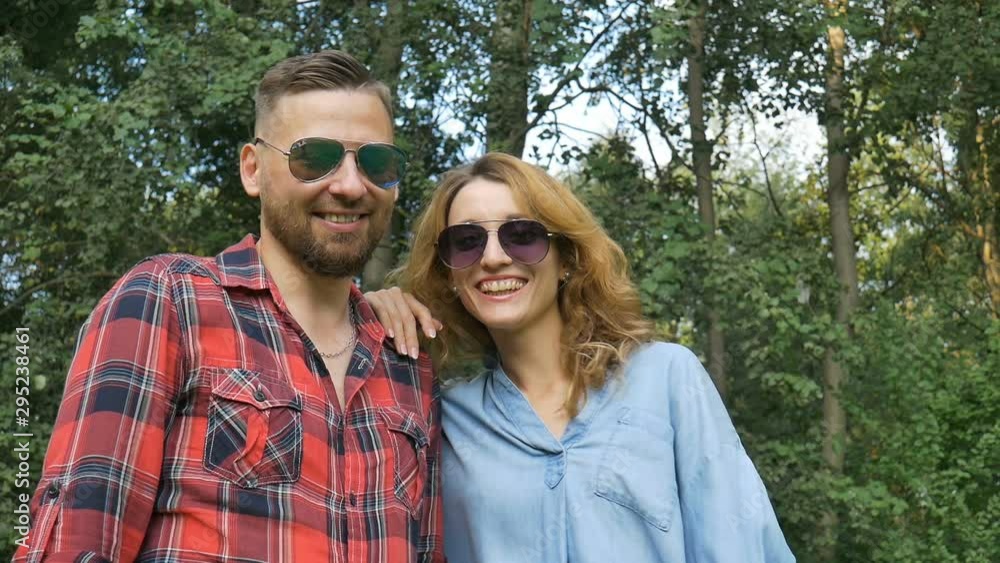 Lovely couple of bearded hipster boyfriend in red shirt and blond girlfriend in blue casual jeans blouse in dark sunglasses looking at the camera and smiling on green trees background Lovely couple of bearded hipster boyfriend in red shirt and blond girlfriend in blue casual jeans blouse in dark sunglasses looking at the camera and smiling on green trees background