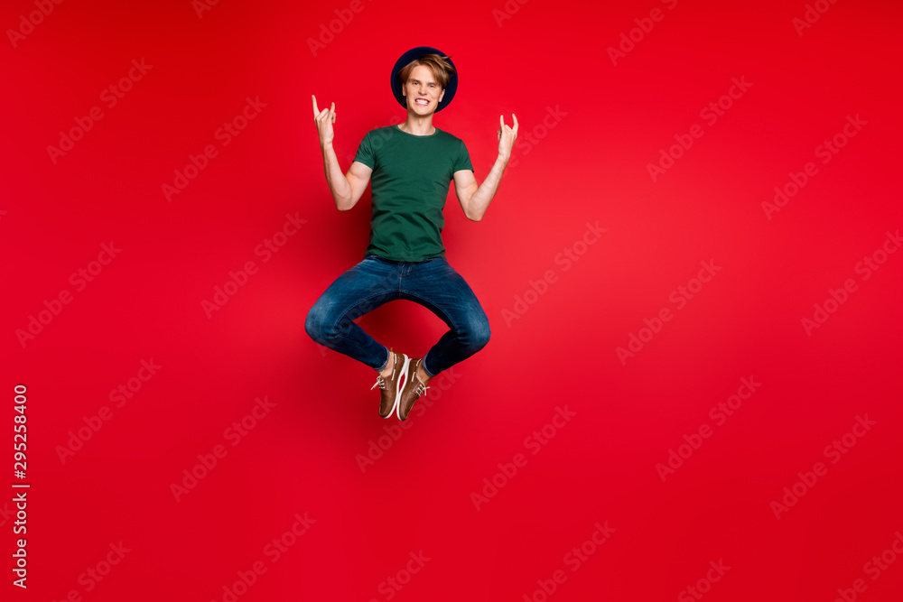 Full length photo of funny funky guy jump show horned sign enjoy rock concert feel candid content crazy wear headwear green t-shirt denim jeans sneakers isolated over red color background Full length photo of funny funky guy jump show horned sign enjoy rock concert feel candid content crazy wear headwear green t-shirt denim jeans sneakers isolated over red color background