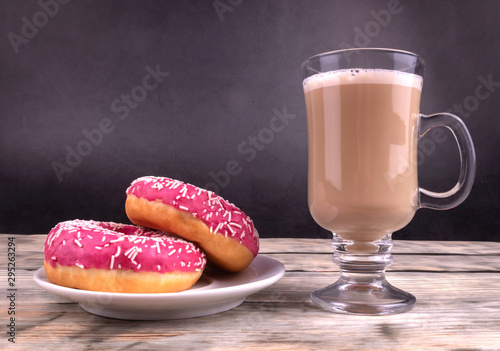 A composition of two pink glazed donuts and a glass cup of coffee with milk