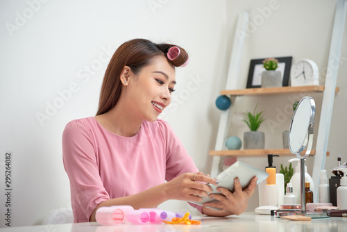 Happy asian woman wearing hair curlers using tablet at home