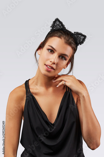 Cropped upper body's shot of a dark-haired girl, posing on a grey background. She is wearing black v-neck tank top and lace kitten ears headband on her head. Her left hand is near face. photo