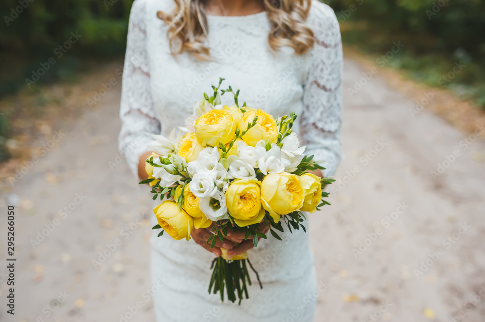 A caucasian woman is standing on pathway, holding a bridal bouquet of yellow roses wearing a wedding dress.
