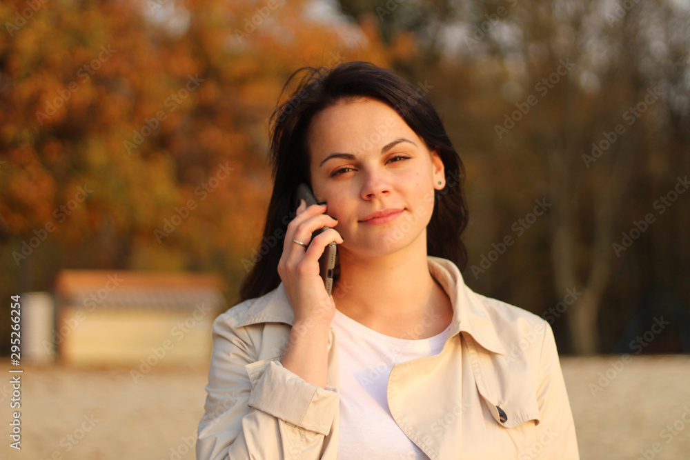 beautiful caucasian girl with dark hair in a raincoat stands on a background of autumn trees holding a smartphone in her hand beautiful caucasian girl with dark hair in a raincoat stands on a background of autumn trees holding a smartphone in her hand