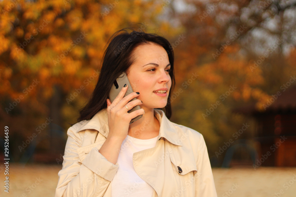 beautiful caucasian girl with dark hair in a raincoat stands on a background of autumn trees holding a smartphone in her hand beautiful caucasian girl with dark hair in a raincoat stands on a background of autumn trees holding a smartphone in her hand