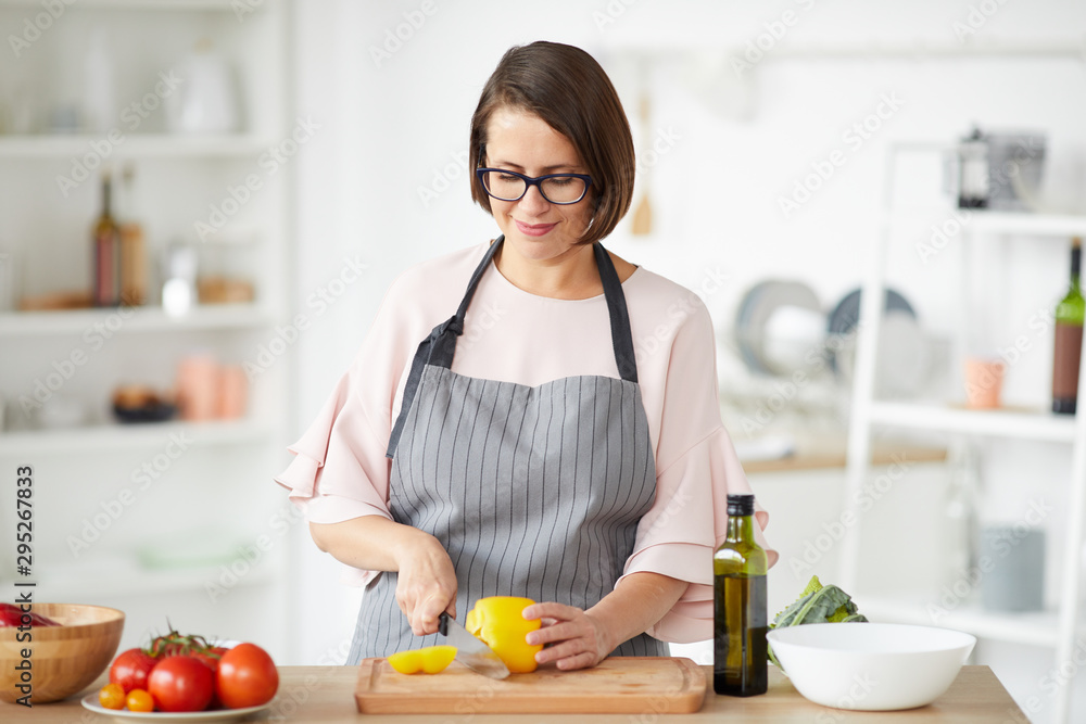 Young housewife in apron standing at the kitchen table and cutting vegetables for salad in the kitchen