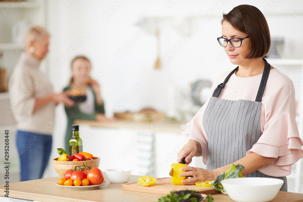 Young woman in eyeglasses wearing apron cutting vegetables on cutting board for dinner while standing in the kitchen with her family in the background