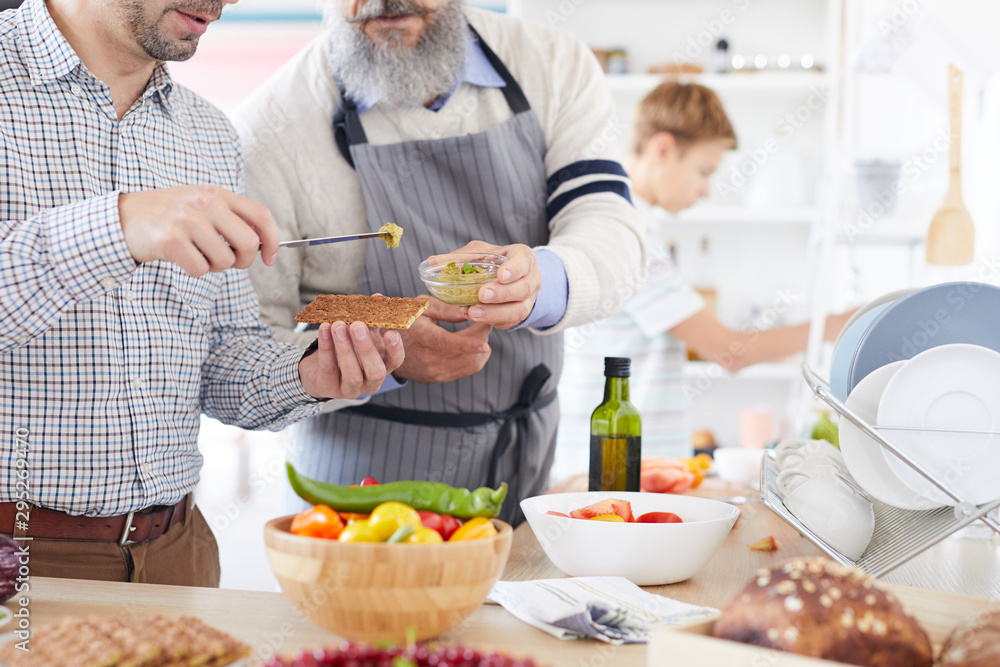 Close-up of young man standing and tasting sauce giving by the mature man while they preparing food in the kitchen Close-up of young man standing and tasting sauce giving by the mature man while they preparing food in the kitchen