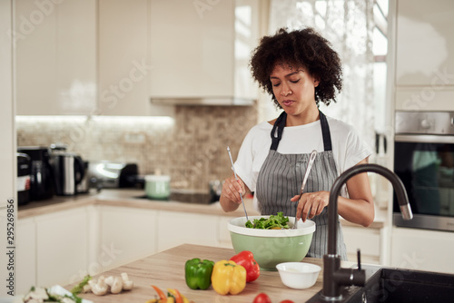 Attractive mixed race woman in apron mixing vegetables in bowl while standing in kitchen at home.