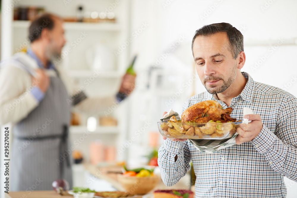 Young man holding dish of roast turkey prepared for holiday with senior man cooking in the background in domestic kitchen Young man holding dish of roast turkey prepared for holiday with senior man cooking in the background in domestic kitchen