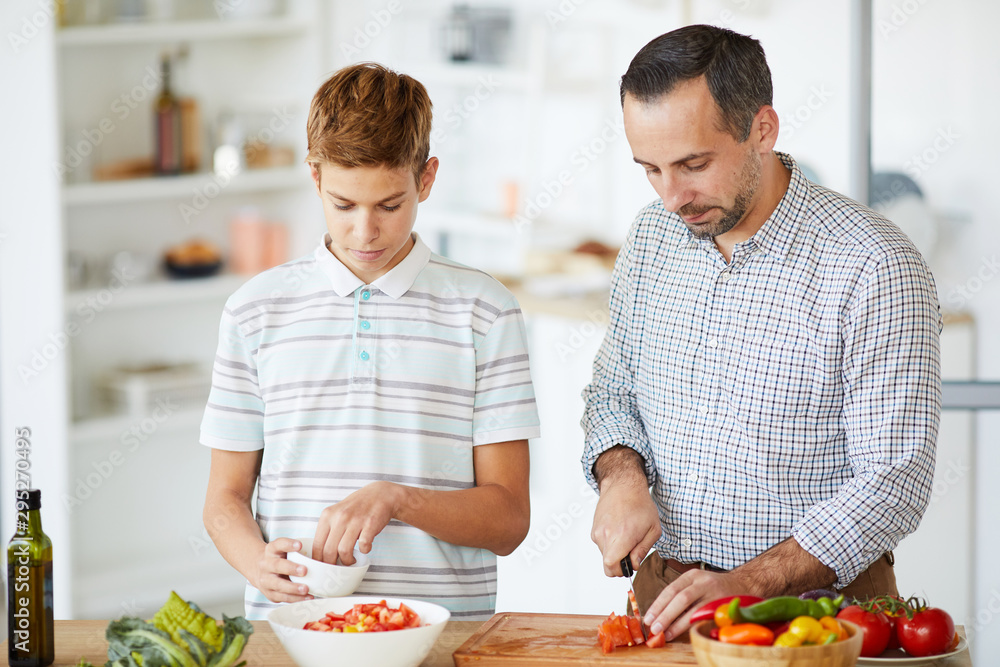 Young father cutting vegetables for salad with his son helping him while they standing at the table in the kitchen