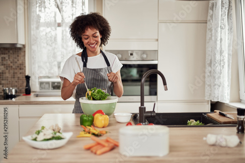 Attractive mixed race woman in apron mixing vegetables in bowl while standing in kitchen at home.