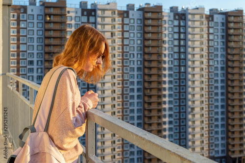 Tween redhead girl in pullover, jeans and sunglasses standing on balcony against high-rise multi-storey residential building at sunset. Beautiful look, fashionable city street outfit, teenage fashion © Tatyana_Andreyeva