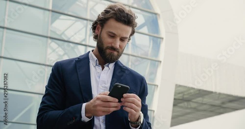 Handsome Businessman browsing his Smartphone near modern Office Building. Attractive Man is smiling and wearing Smart-casual Style. Social Networking. Luxury Lifestyle. Smartphones. Apps.