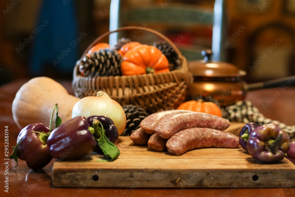 Selective focus still life of preparation of German bratwurst purple peppers and onions on wooden cutting board with a large knife, with blurred basket of pumpkins and pine cones in background Selective focus still life of preparation of German bratwurst purple peppers and onions on wooden cutting board with a large knife, with blurred basket of pumpkins and pine cones in background