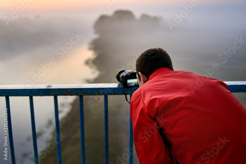 A man on a bridge over the river that photographs a sunrise on a foggy morning.
